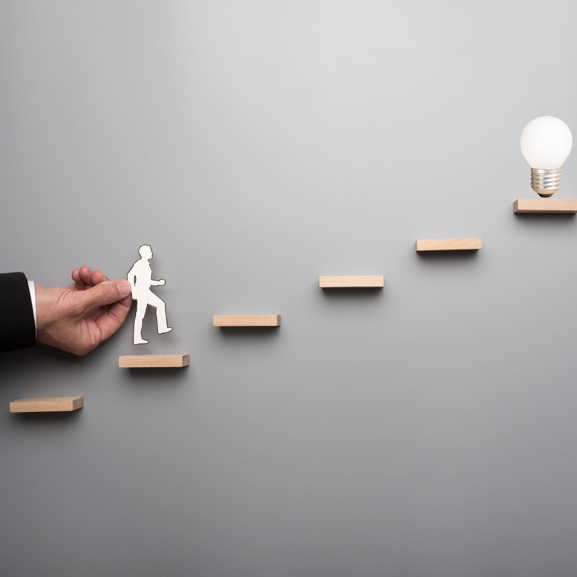 一張灰色背景的圖片,左側有一隻手伸入畫面,拿著一個白色紙片剪影,剪影為一個正在向前行走的人形。這個人形正要踏上一個由五塊木頭磚塊組成的階梯,而階梯的頂端放著一個正在發光的燈泡。